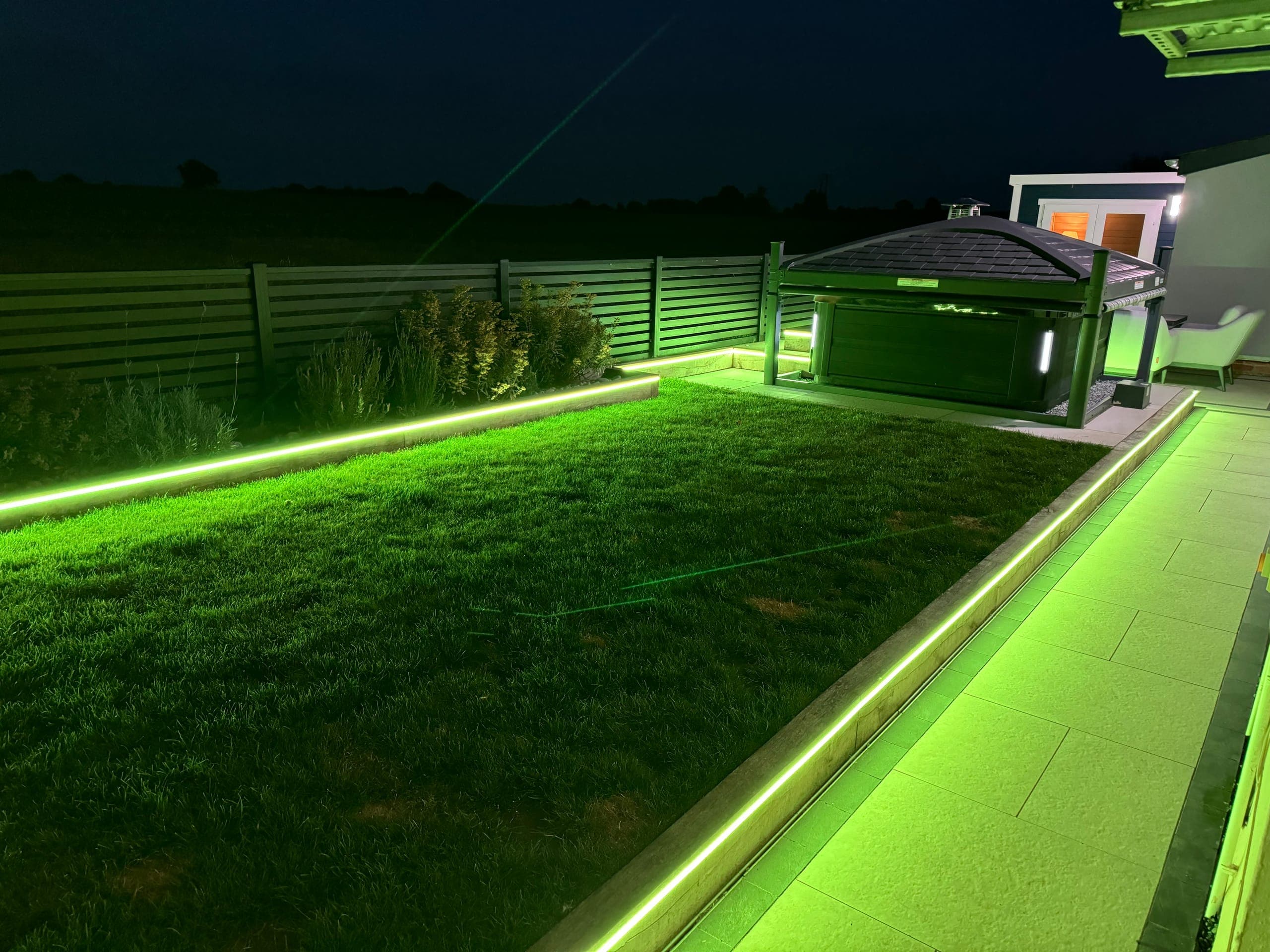Garden lit at night by green LED strip lighting run along raised sod edging and a patio kerb, with a hot tub under a gazebo