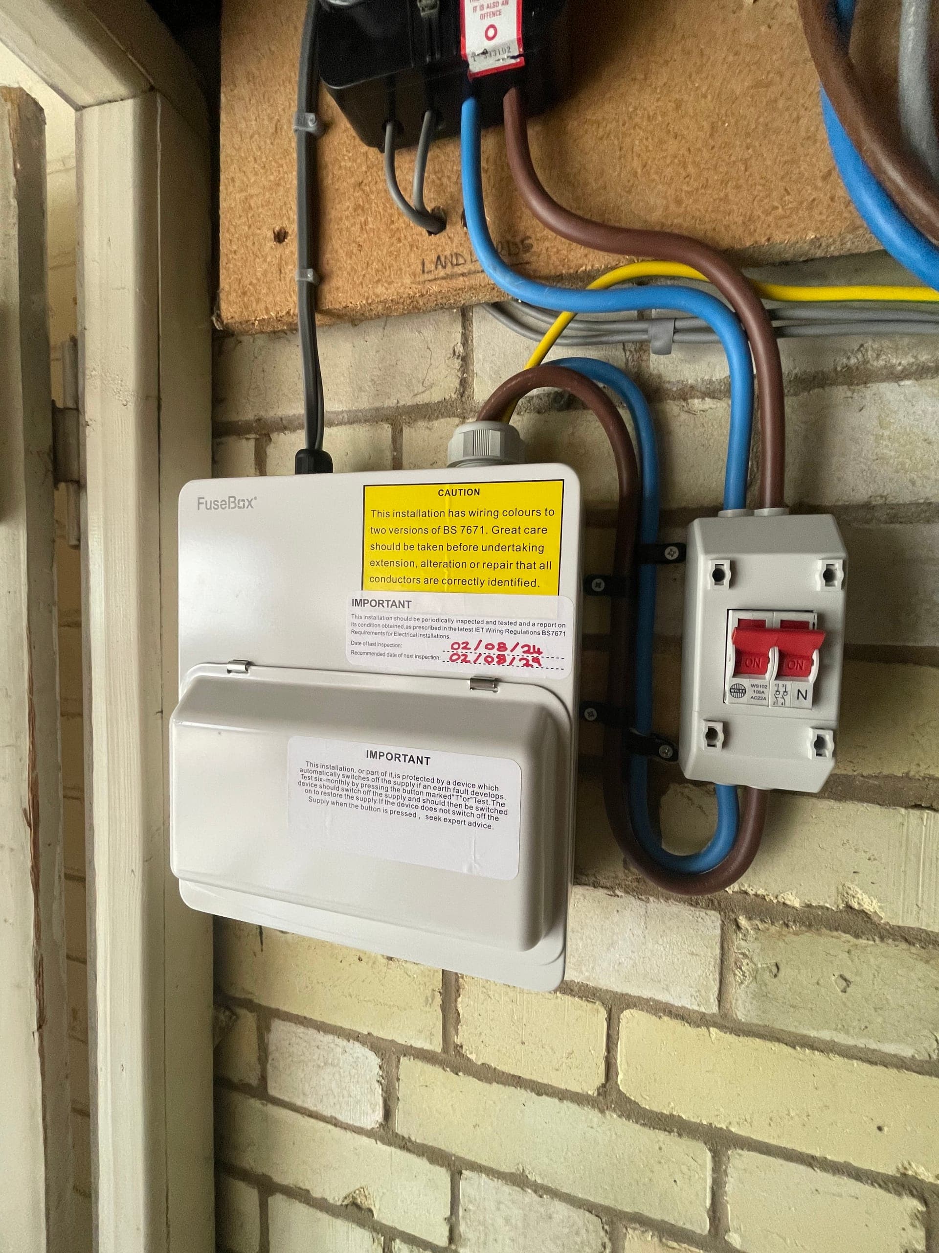 FuseBox consumer unit and 100A double-pole isolator mounted on a brick wall, with inspection labels filled in