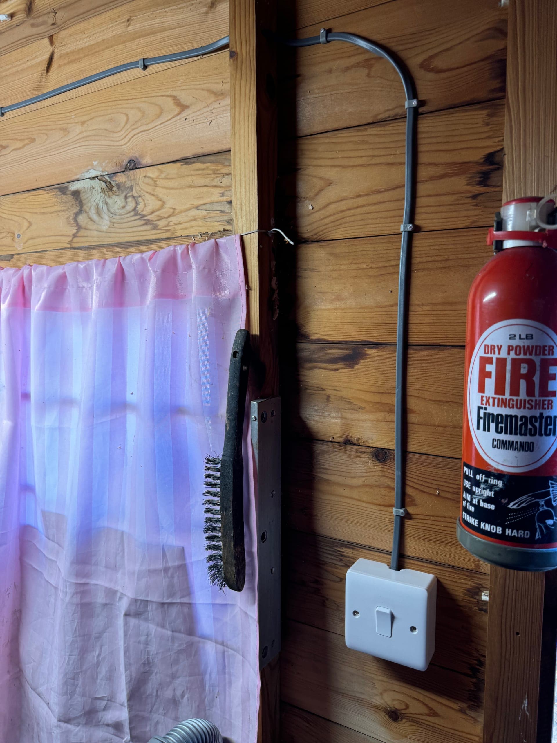 Light switch and surface-clipped cable in a timber outbuilding, beside a fire extinguisher
