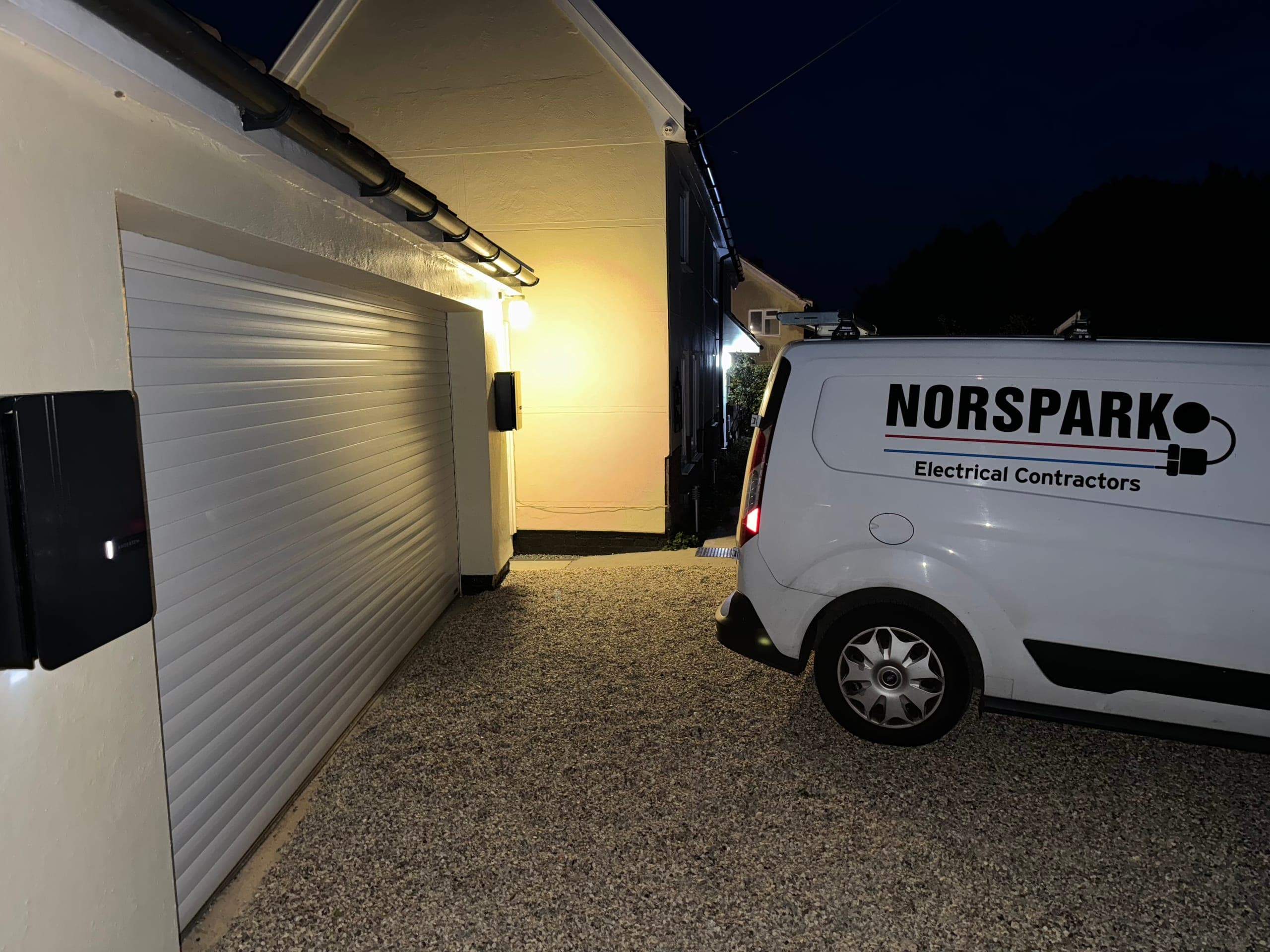 Norspark van on a gravel driveway at night beside a garage, with an outside wall light and a wall-mounted charge point