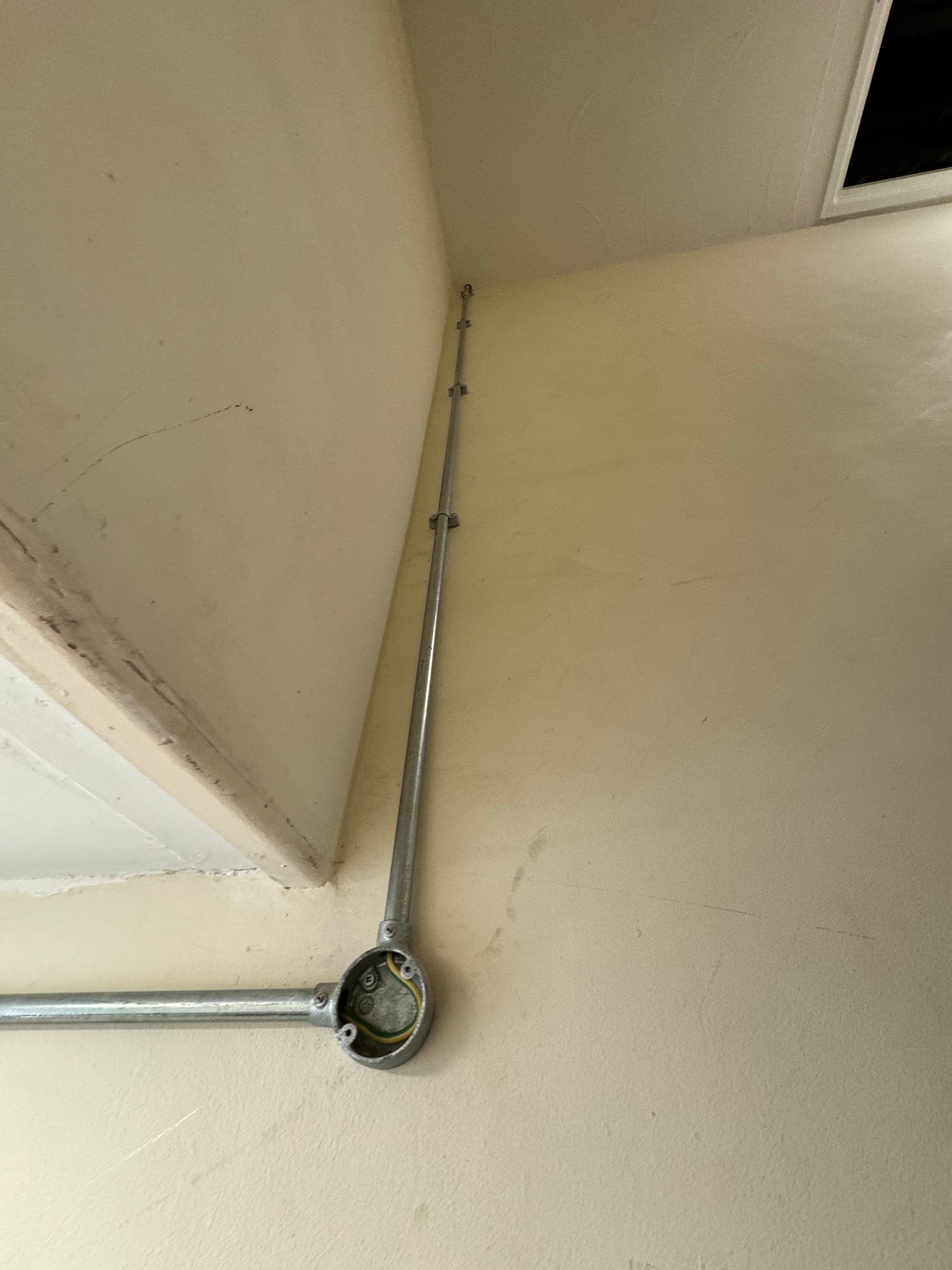 Galvanised conduit meeting a circular junction box in a room corner, earth sleeving visible inside